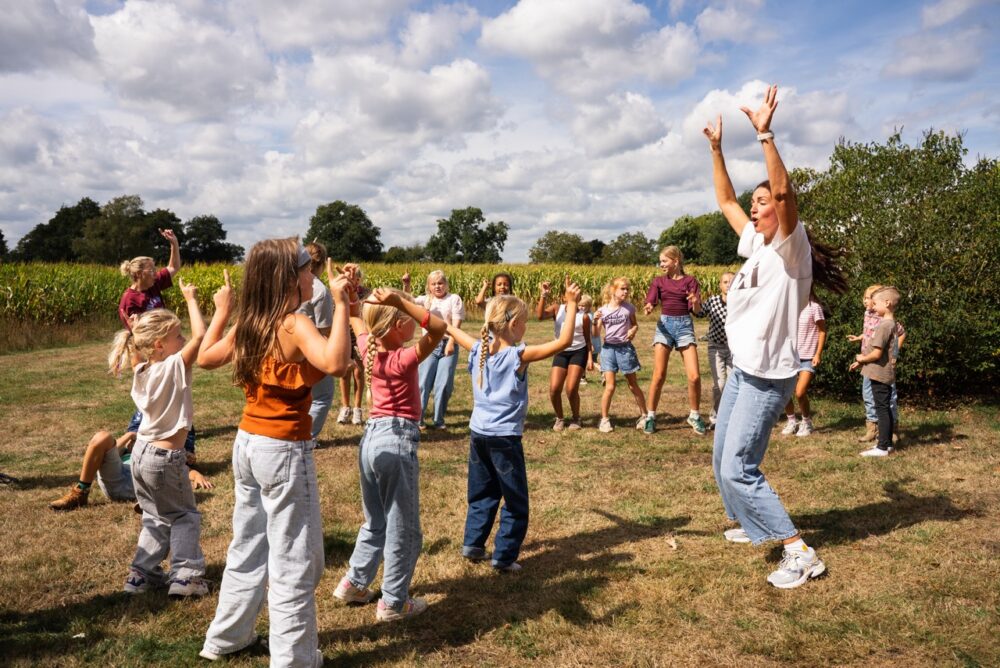 Grote groep kinderen doet met een begeleider een spel op het grasveld tijdens de Kidsdag van Mozaiek0548