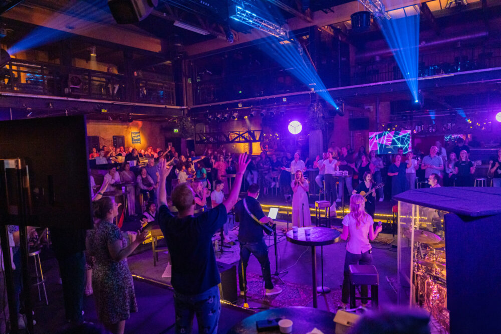 Bezoeker met opgeheven handen op het podium van Mozaiek0548, met de zaal en band eromheen